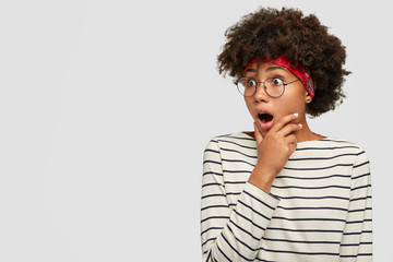 Surprised dark skinned woman keeps jaw dropped, holds chin, looks with astonished expression aside,...