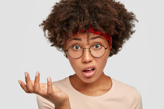 Close up shot of puzzled clueless African girl gestures with puzzlement, looks directly at camera, wears transparent glasses, has dark curly hair, stares with bewilderment, cant understand something