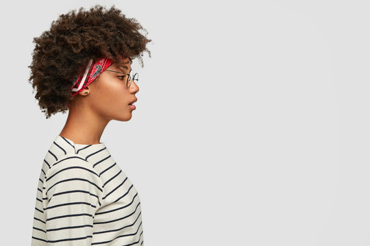 Profile shot of black woman with Afro haircut, thoughtful expression wears headband, round glasses, striped sweater, poses against white background with copy space for your promotional content