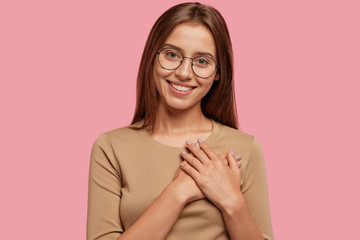 Generous thankful friendly looking girl keeps hands on heart, has good attitude, dressed in casual clothes, isolated over pink background. Joyful kind lady feels thankfulness. Horizontal view
