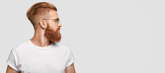 Sideways shot of brutal man with ginger thick beard, trendy hairstyle, looks thoughtfully aside, wears round spectacles and casual t shirt, stands over blank studio background with free space - obrazy, fototapety, plakaty