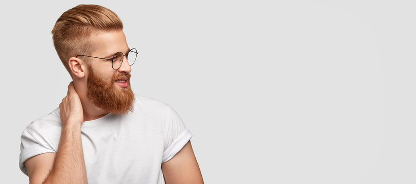 Studio shot of satisfied bearded man with trendy haircut, has charming tender friendly smile, keeps hand on neck, being in good mood, stands against white background with free space for your text
