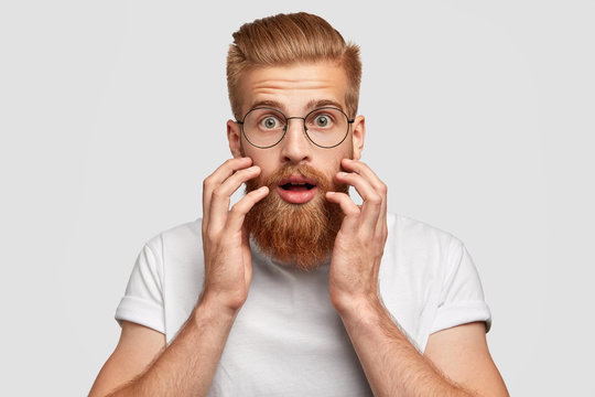 Shocked bearded red haired man looks with stunned expression directly at camera, keeps hands near cheeks, dressed in white t shirt, isolated over white studio wall. People and emotions concept