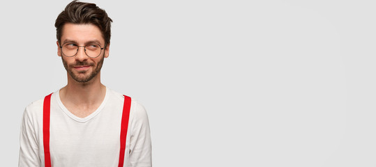 Studio shot of suspicious unshaven man with fashionable hairstyle, round spectacles, looks...