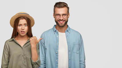 Photo of attractive young girl with puzzled expression points at cheerful guy, express different emotions, stand together against white background with copy space for your advertising content