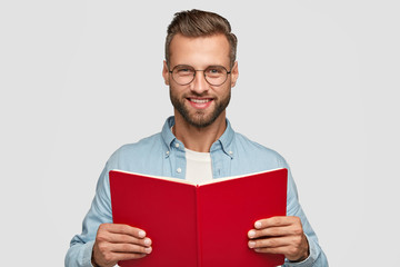 Studio shot of cheerful man reader with satisfied expression, holds red book, reads interesting detective story, wears round transparent glasses for good vision, isolated over white background