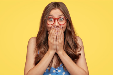 Fototapeta Surprised happy young girl looks with happiness at camera, cant believe in unexpected triumph or success, covers mouth with both hands, wear round spectacles, poses against yellow studio wall obraz