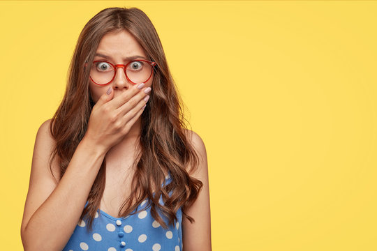 Stupefied attractive European woman covers mouth with palm, has frightened expression as notices something terrible, wears blue polka dot dress, poses against yellow wall, copy space on right