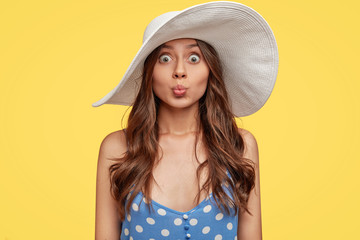 Photo of fashionable attractive woman pouts lips for kiss, wears stylish big white summer hat, polka dor dress, isolated over yellow background, makes grimce, has appealing appearance. Beauty concept © WHstudio Leushin N