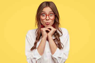European teenager has amazed look at camera, keeps hands under chin, wears spectacles, white shirt, bandana poses over yellow background, pouts lips, has surprised facial expression. Youth concept
