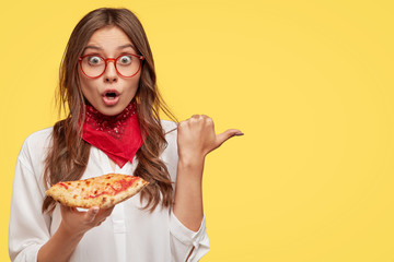 Studio shot of stupefied young female gourmet eats delicious pizza, likes junk food, points aside...