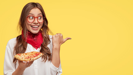 Fastfood and meal concept. Cheerful young girl smiles pleasantly, eats tasty slice of pizza, being...