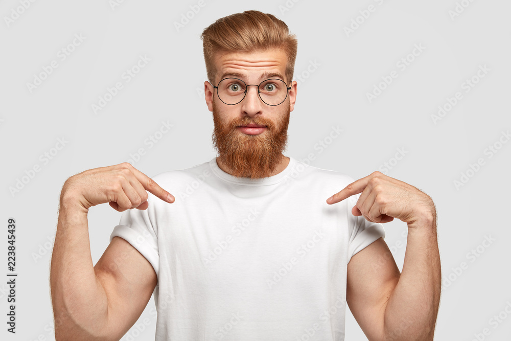 Wall mural stupefied red haired man has thick beard, points at copy space of t shirt, shows place for your slog