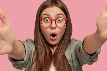 Stupefied Caucasian woman with surprised terrified expression, stretches hands for making selfie,...