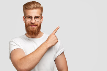 Studio shot of ginger hipster with thick beard, trendy haircut, has serious expression, points with index finger at upper right corner, dressed in white t shirt shows free space for your advertisement