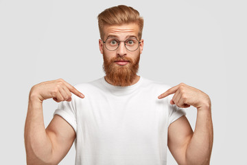 Stupefied red haired man has thick beard, points at copy space of t shirt, shows place for your...