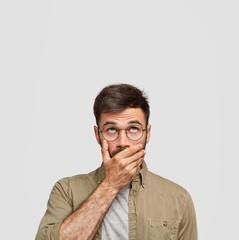 Vertical shot of handsome man with dark hair, keeps hand over mouth, being speechless, dressed in...
