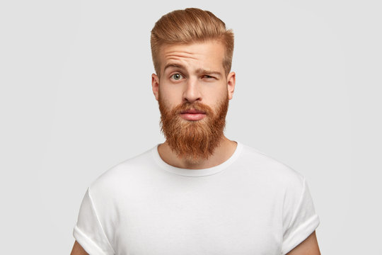 Bewildered man with thick ginger beard, raises eyebrows, reacts on fake news from friend, looks directly at camera, dressed in casual t shirt, isolated over white background. Facial expressions