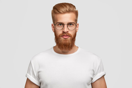 Horizontal shot of attractive hipster looks seriously directly at camera, being self assured, wears big round spectacles, white t shirt in tone with background. Masculinity and style concept