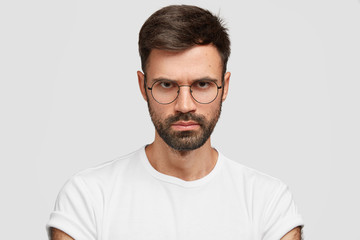 Obraz premium Close up shot of confident strict businessman has thick bristle, looks with gloomy expression, frowns face, wears glasses and casual t shirt, expresses negative feeling, isolated over white background