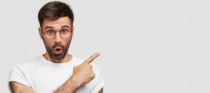 Photo of shocked European man with thick bristle points at blank copy space, being surprised and impressed to see something unbelievable, stands against white background. Look on right side.