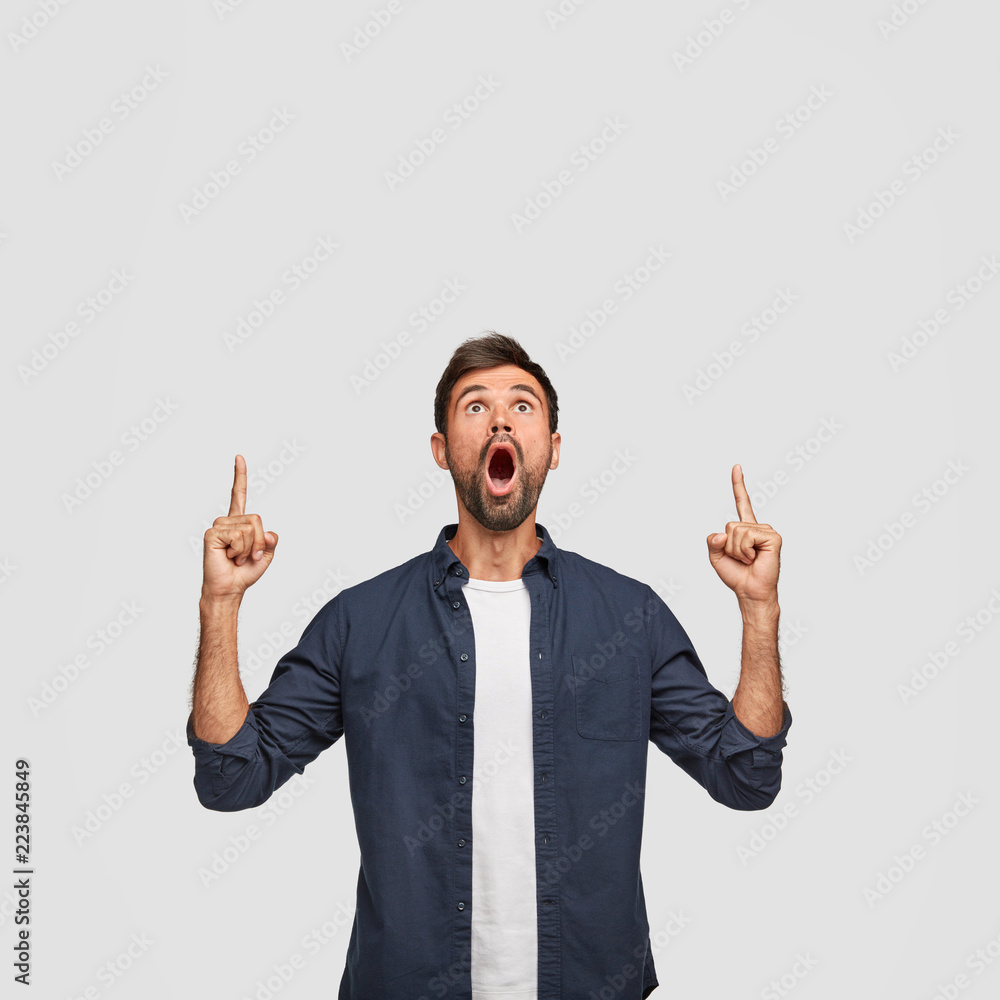 Wall mural Shocked bearded guy with surprised look, points with both index fingers above, wonders as sees something unbelievable, dressed in fashionable shirt, poses against white background. Advertisement