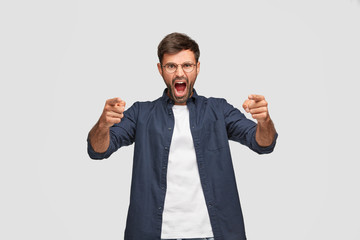 Horizontal shot of angry furious man with annoyed expression, yells at someone, points with both index fingers at camera, blames you about something, poses over white background. Irritated guy indoor