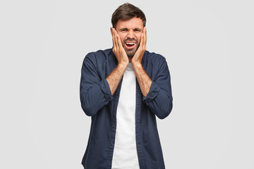 Dejected astonished displeased man frowns face, keeps hands on cheeks, regrets something, has...