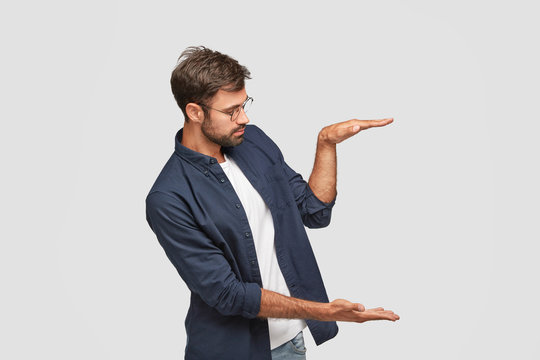 Sideways shot of self assured man demonstrates size or height of something, raises palms in air against white background, wears round spectacles, isolated over white background. Body language concept