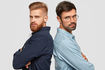 Two companions stand back to each other, look seriously at camera, keep arms folded, work as team,...