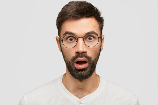 Headshot of stunned young European guy with thick stubble, stares at camera, hears unexpected rumours, wears casual clothes, poses against white background. People and facial expressions concept
