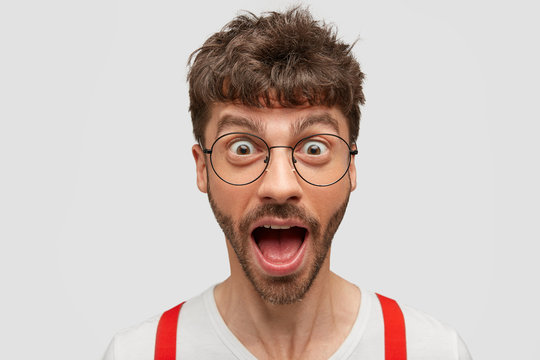 Horizontal shot of funny young man opens mouth with surprisement, popped eyes, wears round transparent glasses, thrilled to recieve awesome news, poses over white background. Emotions concept
