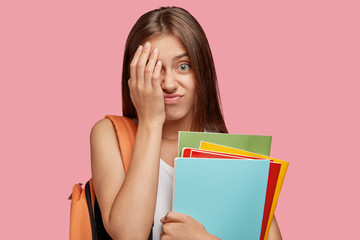 Displeased European teenager covers face with frustrated expression, feels tired after preparing diploma paper, carries textbooks, rucksack, poses against pink studio wall. Overworked pupil.