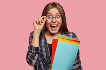 Amazed young European girl opens mouth from amazement, keeps hand on rim of spectacles, dressed in...