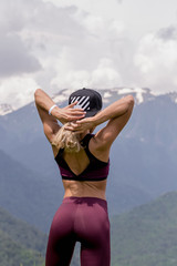 Young sportive blonde female trainer in active wear exercising outdoors, looking at panoramic view of mountain range in the mist under the sunrise.