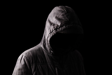 Scary and creepy man hiding in the shadows, with the face and identity hidden with the hood, and standing in the darkness. Low key, black background. Concept for fear, mystery, danger, crime, stalker