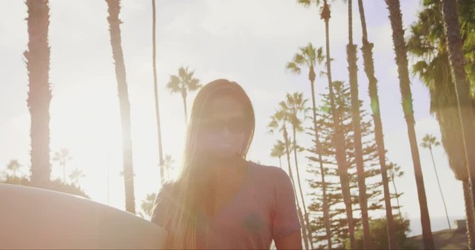Close up of attractive surfer girl walking with skateboard down street at sunset with lens flare 