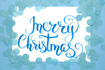 Christmas flat lay: lettering text 'Merry Christmas' andblue confetti on blue background.