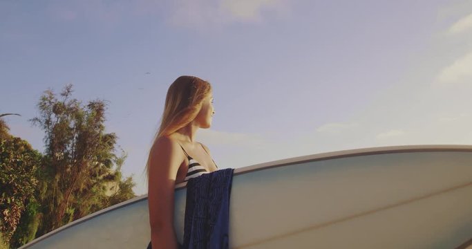 Happy surfer girl looking out to ocean smiling with surfboard under her arm 