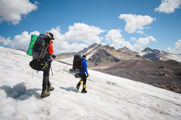Two tourists, a man and a woman with backpacks and crampons on their feet walk along the glacier against the background of the mountains of the sky and clouds. Back view