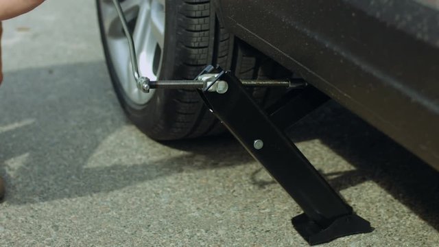 Close-up of male hand lifting car with spanner jack-screw to change flat tyre on road after puncture. Man jacking up auto with car jack to replace tire during road trip.