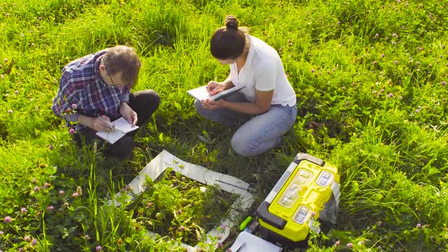 Crane shot. Two ecologist examining plants on the meadow. Woman taking protective gloves from toolbox. Field work. High angle view