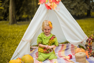 Fototapeta premium Little girl lying and playing in a tent, children's house wigwam in park Autumn portrait of cute curly girl. Harvest or Thanksgiving. autumn decor, party, picnic. Child in halloween costume.