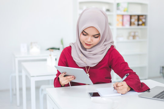 Young Asian Muslim Woman Using Tablet And Taking Notes In Notebook In A White Cafe.