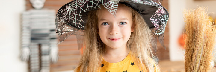 Cute little girl in witch costume holding a broom is standing in Halloween decorated living room,...