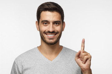 Fototapeta premium Handsome smiling young male in gray t-shirt pointing up with his finger, isolated on grey background