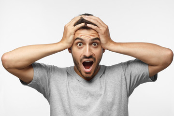 Emotional young man with strong expression of fear, round eyes, open mouth and hands pressed to top of head, showing astonishment and shock, isolated on gray background