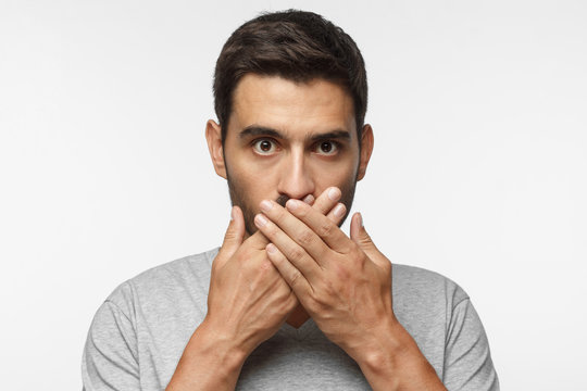 Secret, silence concept. Close up portrait of young man in gray t-shirt covering his mouth with hands isolated on grey background