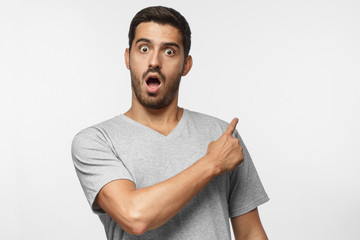 Young surprised man pointing right, isolated on gray background in gray t-shirt, looking at camera...