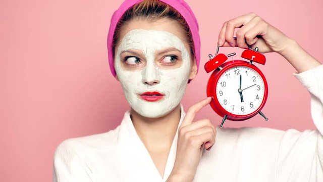 A girl with a mask on her face and a towel on her head holds an alarm clock in her hand on a pink background. Face care concept. Beautiful girl with cosmetic mask on her face.